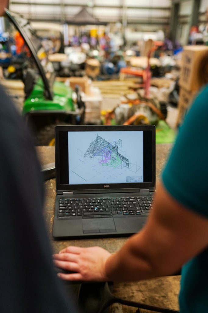 Two engineers reviewing architectural plans on a laptop in a busy workshop.