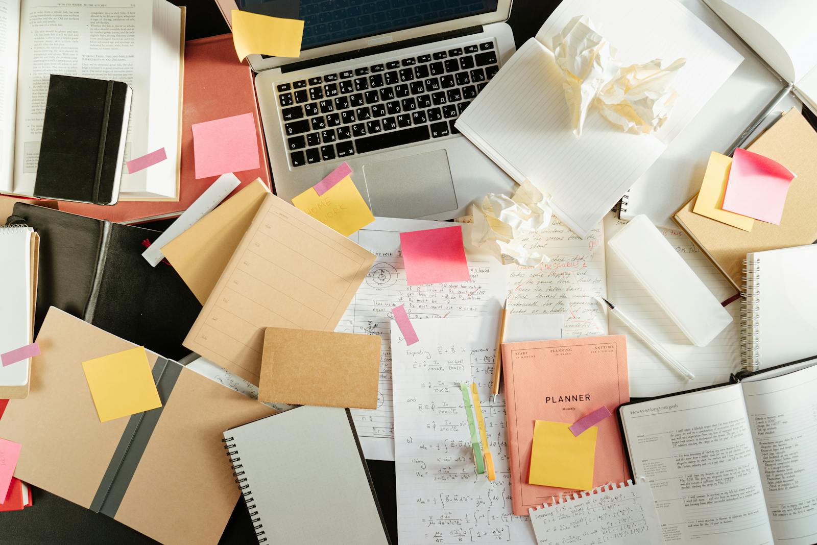 Flatlay of a messy study desk with laptop, notebooks, and sticky notes, showcasing the creative process.