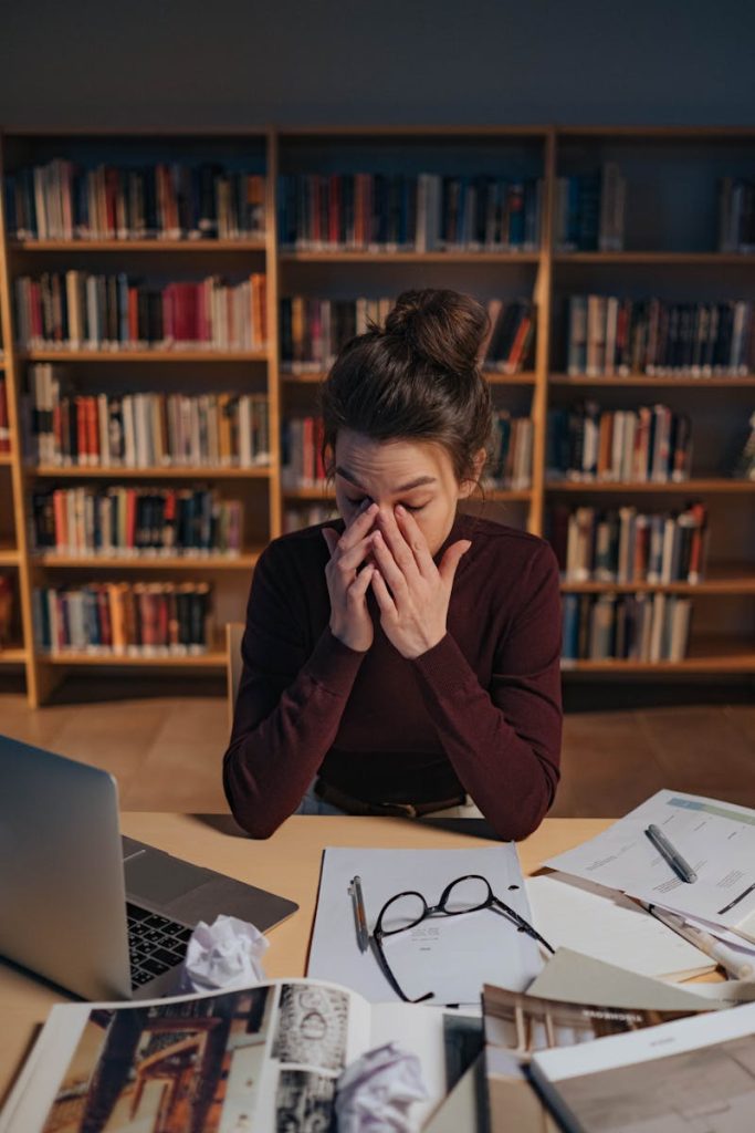 Woman feeling stressed while studying in a library, surrounded by books and laptop.