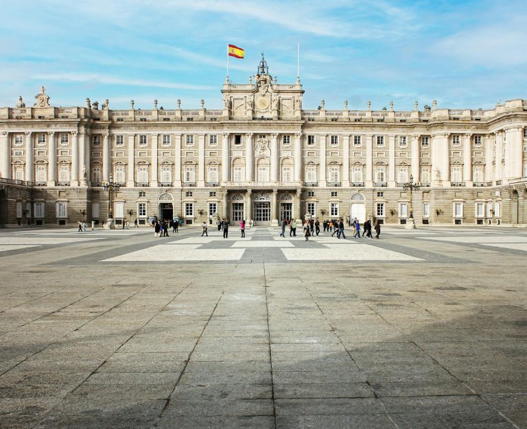 A view of the Royal Palace of Madrid with tourists in the courtyard under a sunny sky.