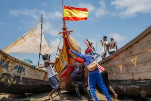 Group of men pushing colorful fishing boats on the shore with the Spanish flag flying high.